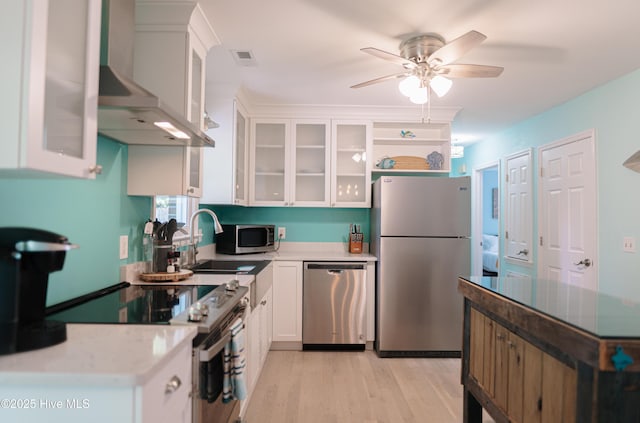 kitchen with appliances with stainless steel finishes, wall chimney exhaust hood, a ceiling fan, a sink, and white cabinets