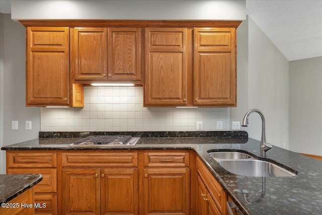 kitchen with a sink, stainless steel gas cooktop, dark stone counters, backsplash, and brown cabinets