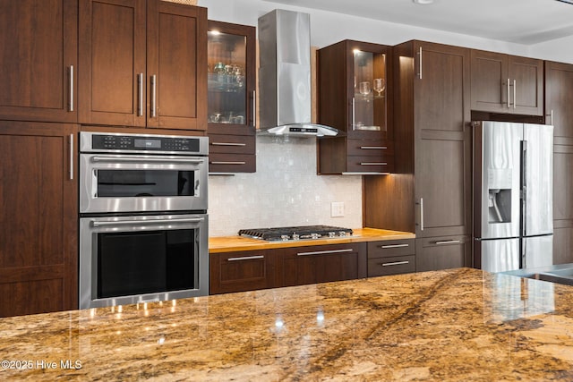 4145 Brighton Ln, Southport, NC 28461 - kitchen with stainless steel appliances, wall chimney range hood, light stone counters, backsplash, and glass insert cabinets