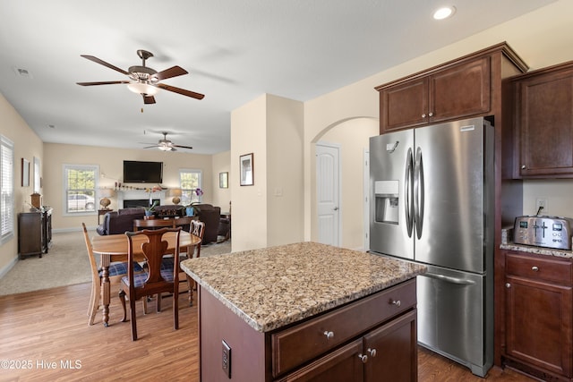 kitchen featuring stainless steel refrigerator with ice dispenser, ceiling fan, wood finished floors, a kitchen island, and light stone countertops