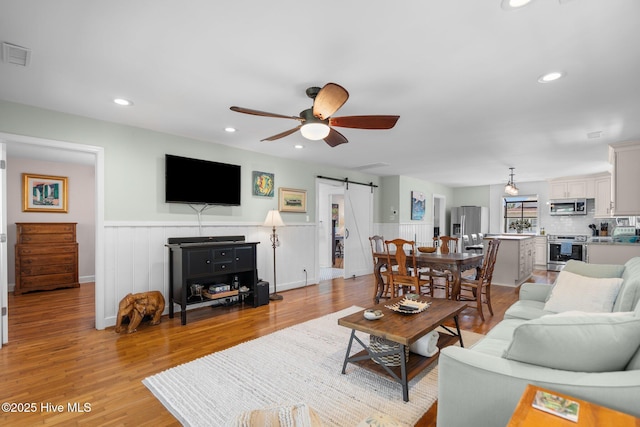 living room featuring a barn door, a ceiling fan, light wood-style flooring, a wainscoted wall, and recessed lighting