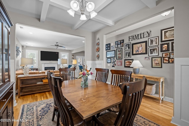 dining room featuring light wood finished floors, a fireplace, beam ceiling, and ceiling fan with notable chandelier