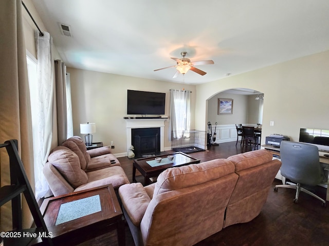 living room featuring visible vents, ceiling fan, arched walkways, dark wood-style floors, and a fireplace with flush hearth