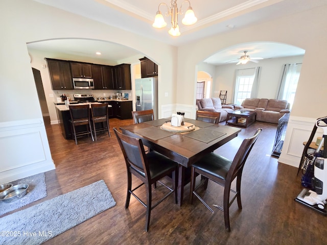 dining room featuring ceiling fan with notable chandelier, ornamental molding, dark wood finished floors, arched walkways, and wainscoting