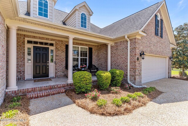 entrance to property with a shingled roof, a porch, driveway, brick siding, and an attached garage