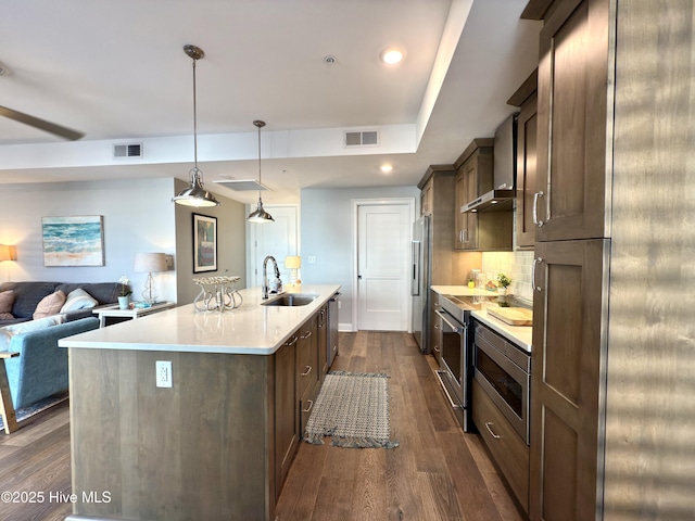 kitchen with stainless steel appliances, a sink, wall chimney exhaust hood, dark wood-type flooring, and light countertops