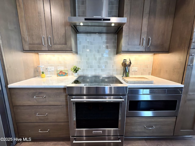 kitchen with wall chimney range hood, tasteful backsplash, light countertops, dark brown cabinets, and appliances with stainless steel finishes