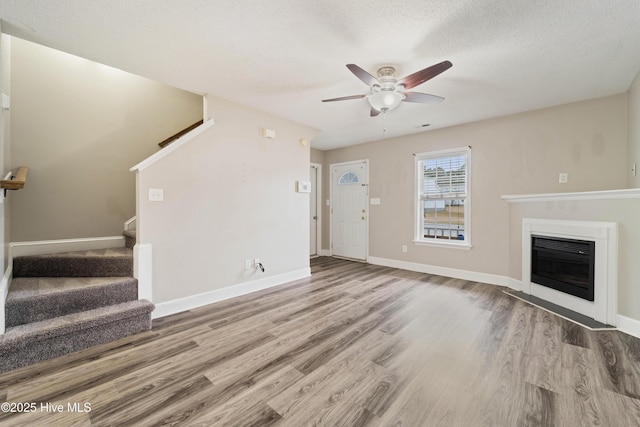 unfurnished living room featuring wood finished floors, stairway, baseboards, ceiling fan, and a glass covered fireplace