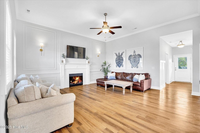 living area with a decorative wall, a warm lit fireplace, ornamental molding, ceiling fan, and light wood-style floors