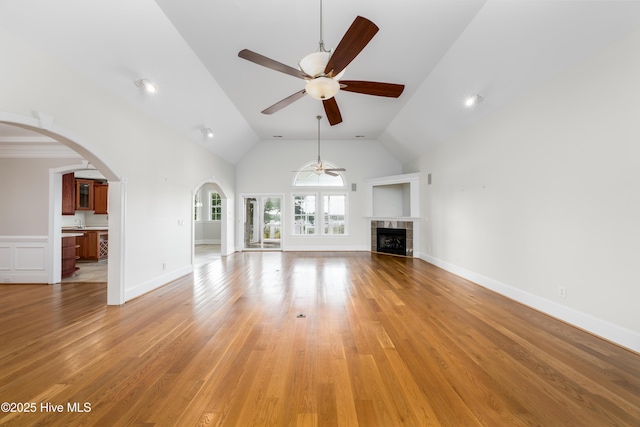 unfurnished living room with a ceiling fan, arched walkways, a tile fireplace, light wood-style floors, and high vaulted ceiling