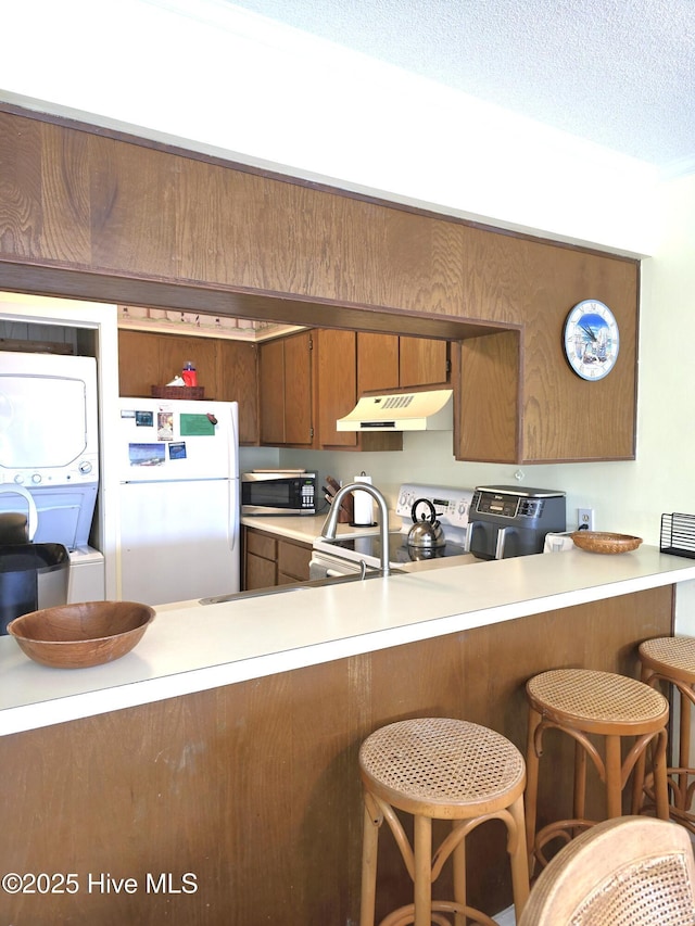 kitchen featuring stacked washer / dryer, under cabinet range hood, white appliances, brown cabinetry, and light countertops