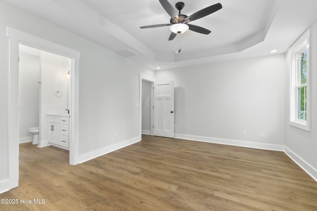 unfurnished bedroom with wood finished floors, a tray ceiling, baseboards, ensuite bath, and recessed lighting