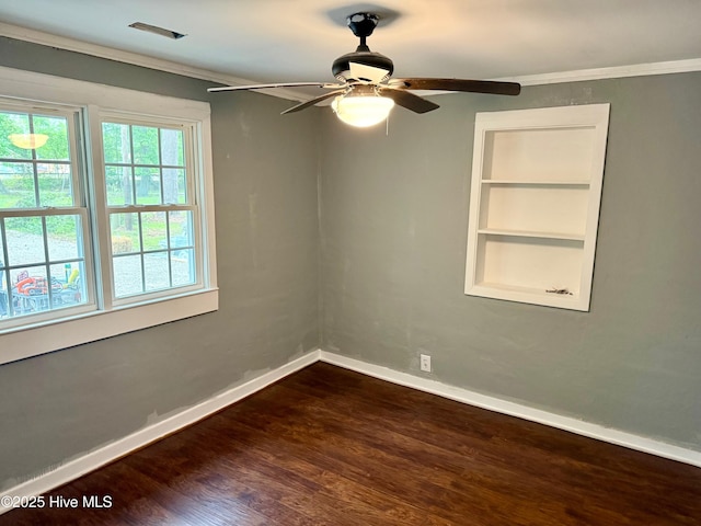 empty room featuring dark wood-type flooring, built in shelves, crown molding, a ceiling fan, and baseboards
