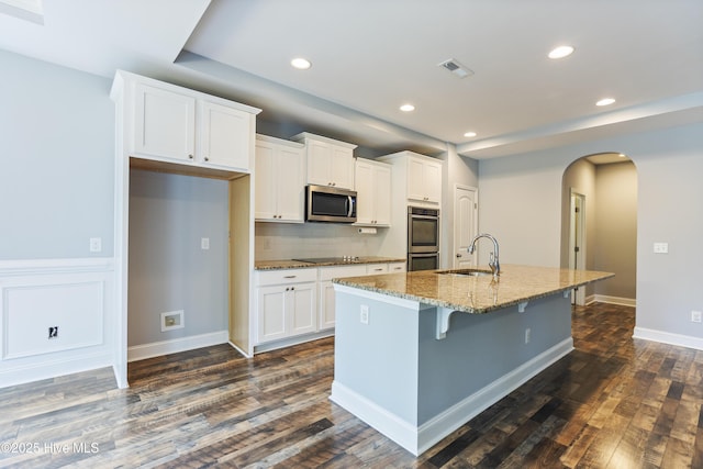kitchen featuring appliances with stainless steel finishes, a sink, arched walkways, a kitchen island with sink, and white cabinets