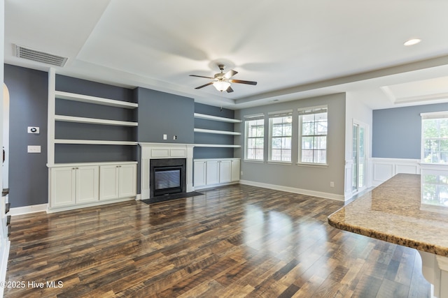 unfurnished living room with a fireplace with flush hearth, a ceiling fan, dark wood-type flooring, built in features, and a raised ceiling
