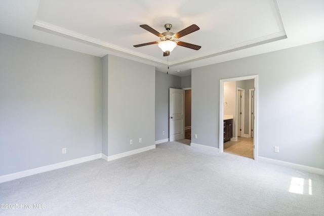 unfurnished bedroom with a tray ceiling, light colored carpet, baseboards, connected bathroom, and a ceiling fan
