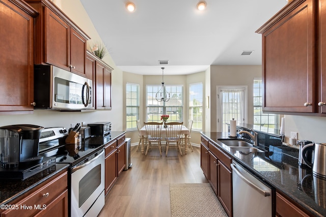 kitchen with appliances with stainless steel finishes, a sink, light wood finished floors, dark stone counters, and hanging light fixtures