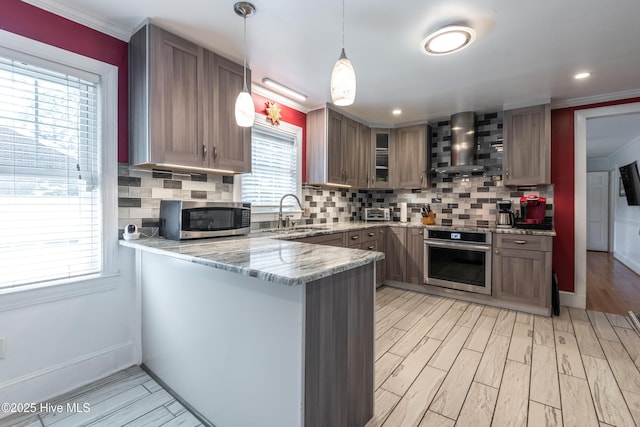 kitchen with wall chimney exhaust hood, appliances with stainless steel finishes, a sink, a peninsula, and light stone countertops