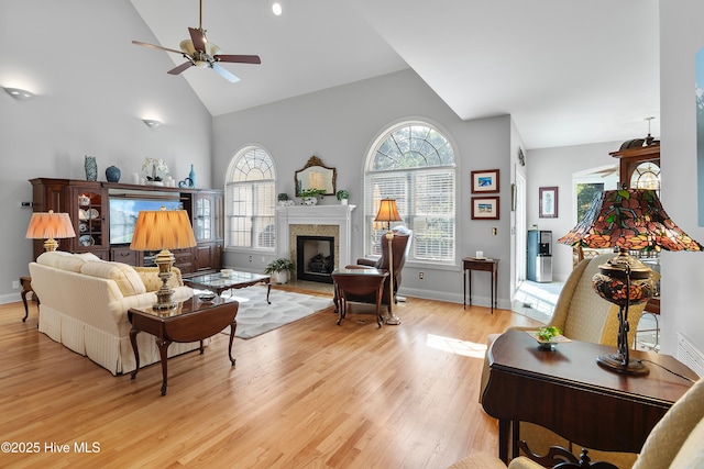 living area with high vaulted ceiling, a fireplace, ceiling fan, light wood-style flooring, and baseboards