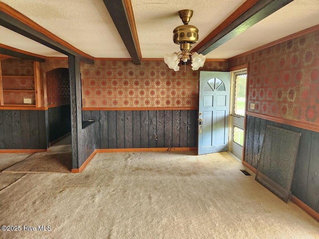 unfurnished dining area with wainscoting, wallpapered walls, carpet flooring, ceiling fan, and beamed ceiling