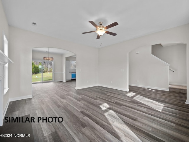 unfurnished living room with arched walkways, a ceiling fan, dark wood-style floors, baseboards, and a chandelier