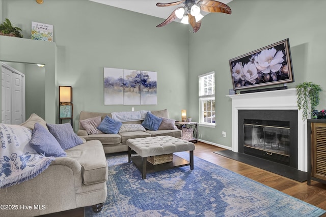 living room featuring a towering ceiling, a ceiling fan, wood finished floors, a glass covered fireplace, and baseboards