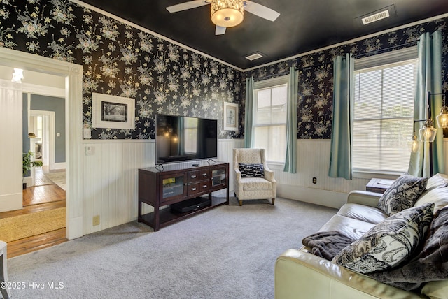 carpeted living room with wainscoting, wallpapered walls, a ceiling fan, and ornamental molding