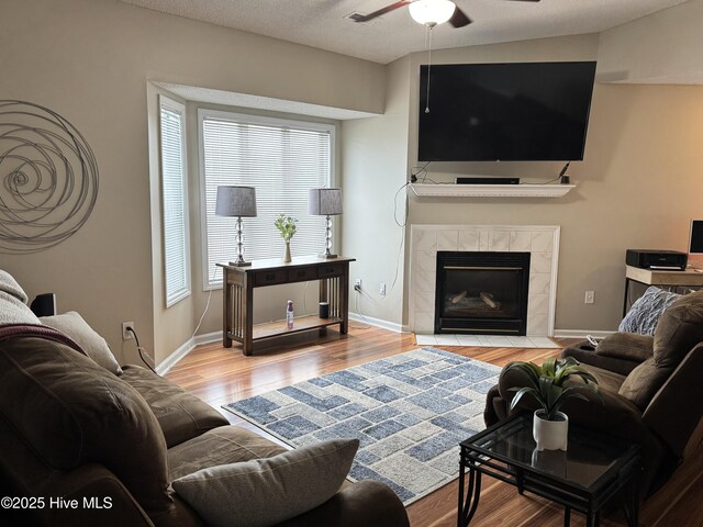 living room with ceiling fan, wood finished floors, a tiled fireplace, and baseboards