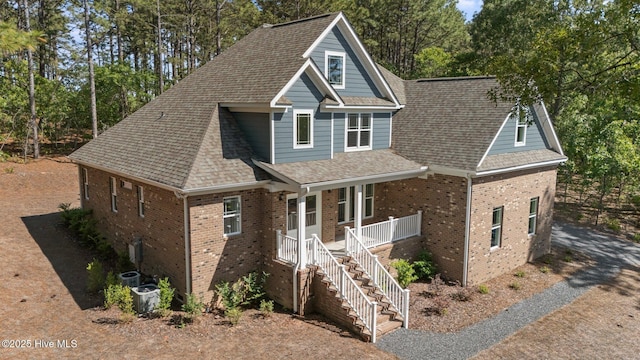 view of front of home featuring covered porch, brick siding, roof with shingles, stairs, and view of wooded area