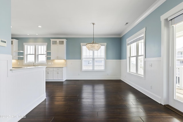 unfurnished dining area featuring a wainscoted wall, a chandelier, plenty of natural light, dark wood-type flooring, and ornamental molding