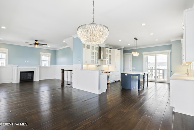kitchen featuring wall chimney range hood, a ceiling fan, white cabinetry, a center island, and crown molding
