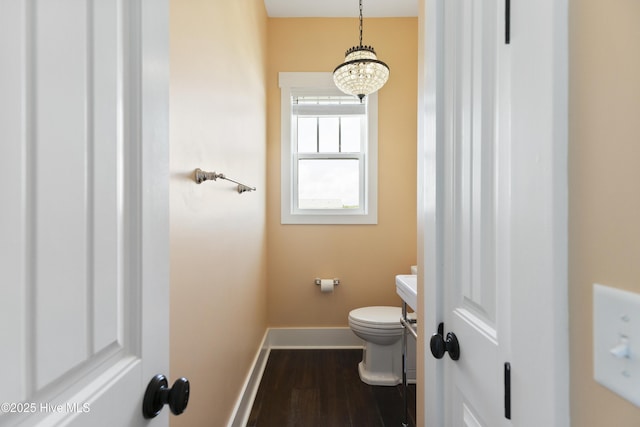 half bath with wood finished floors, baseboards, toilet, vanity, and a chandelier