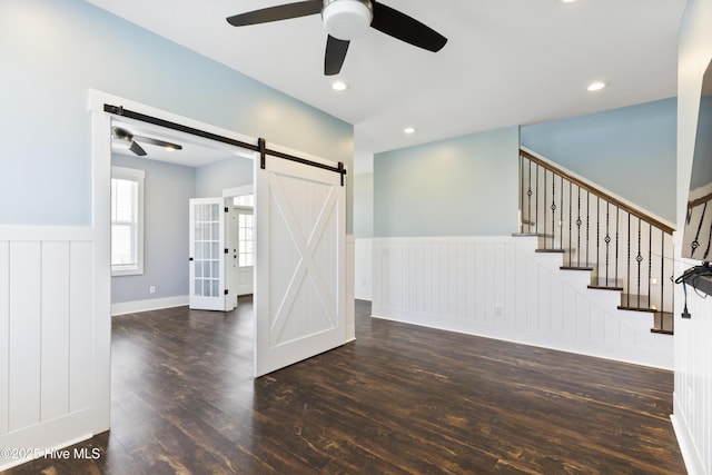 unfurnished living room featuring ceiling fan, wainscoting, wood finished floors, a barn door, and stairway