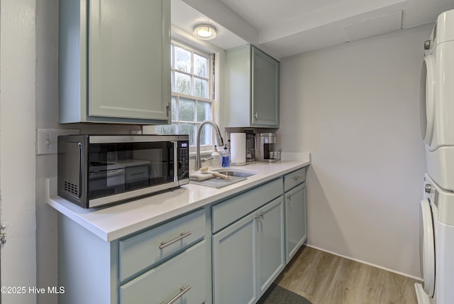kitchen with a sink, stainless steel microwave, stacked washer and clothes dryer, light wood-type flooring, and light countertops