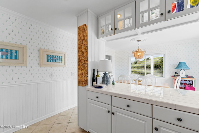 kitchen with wallpapered walls, crown molding, a chandelier, wainscoting, and glass insert cabinets