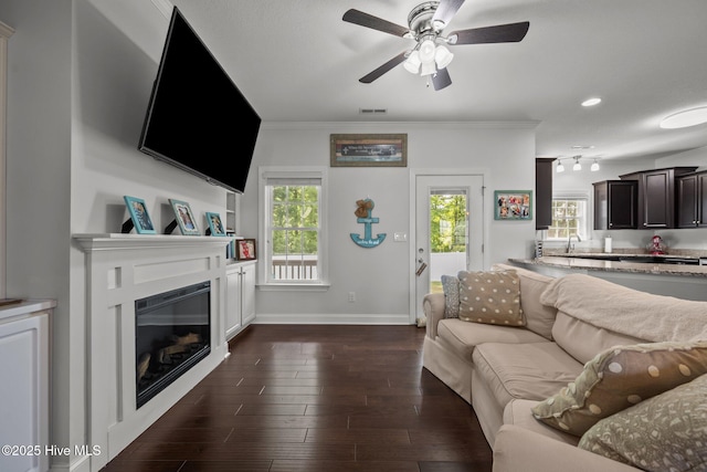 living room featuring a ceiling fan, a glass covered fireplace, baseboards, ornamental molding, and dark wood-style flooring