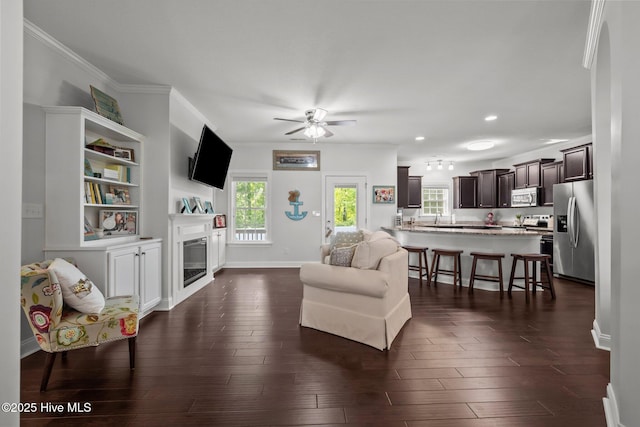 living area featuring a glass covered fireplace, ceiling fan, ornamental molding, recessed lighting, and dark wood-style flooring