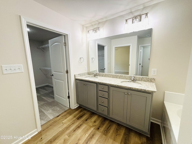full bath featuring a walk in closet, wood finished floors, double vanity, a bathtub, and baseboards