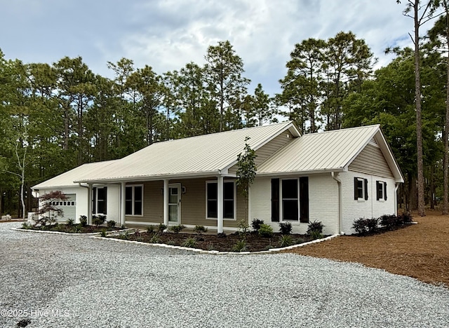 ranch-style house with gravel driveway, metal roof, brick siding, and view of wooded area