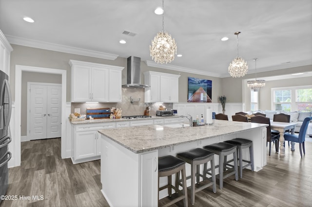 kitchen featuring wall chimney exhaust hood, a chandelier, appliances with stainless steel finishes, crown molding, and white cabinets