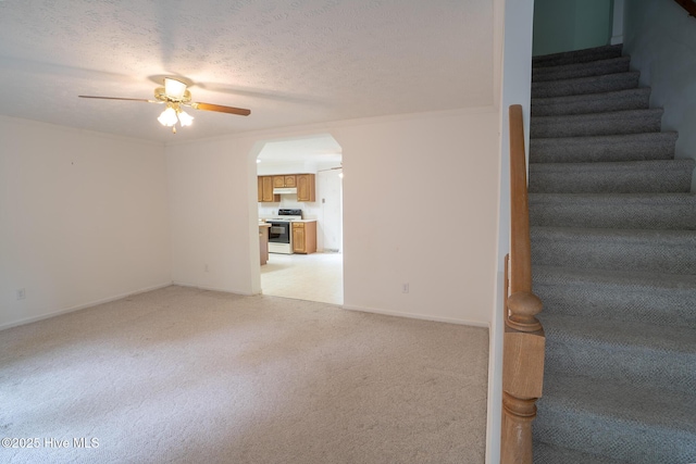 unfurnished living room with light carpet, ceiling fan, arched walkways, stairs, and a textured ceiling
