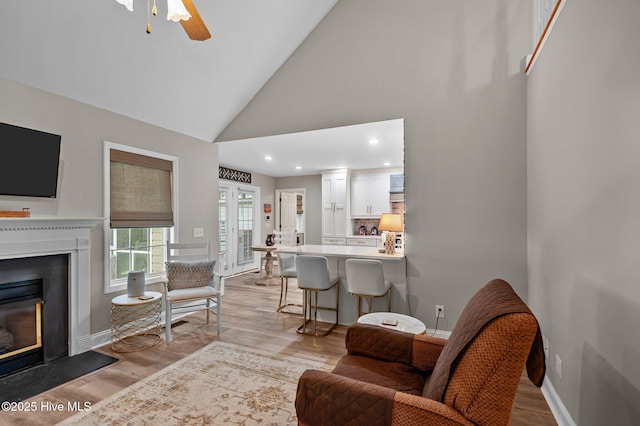 living room with a glass covered fireplace, light wood finished floors, high vaulted ceiling, baseboards, and a ceiling fan