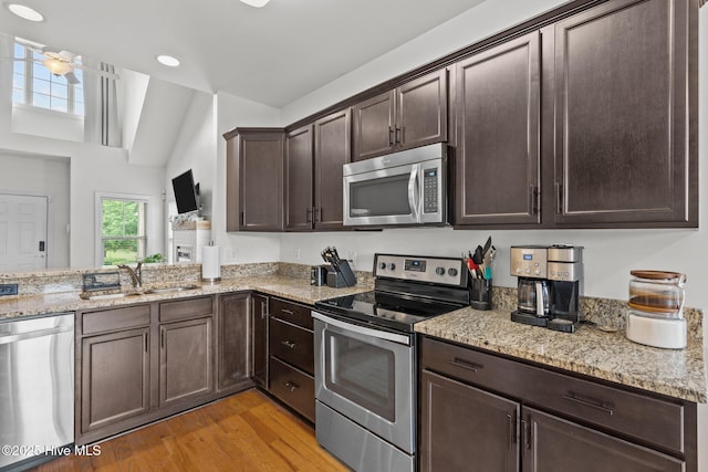 kitchen with appliances with stainless steel finishes, a sink, dark brown cabinetry, light wood finished floors, and light stone counters