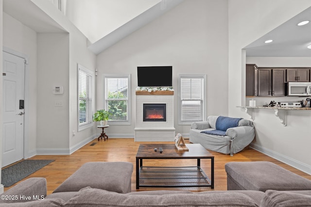 living room with high vaulted ceiling, light wood finished floors, a glass covered fireplace, recessed lighting, and baseboards