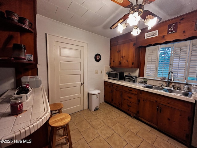 kitchen featuring tile countertops, a sink, stainless steel microwave, ceiling fan, and crown molding