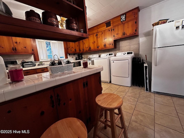 kitchen with freestanding refrigerator, tile counters, light tile patterned floors, independent washer and dryer, and brown cabinets