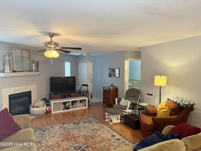 living room with ceiling fan, wood finished floors, a tiled fireplace, and a textured ceiling