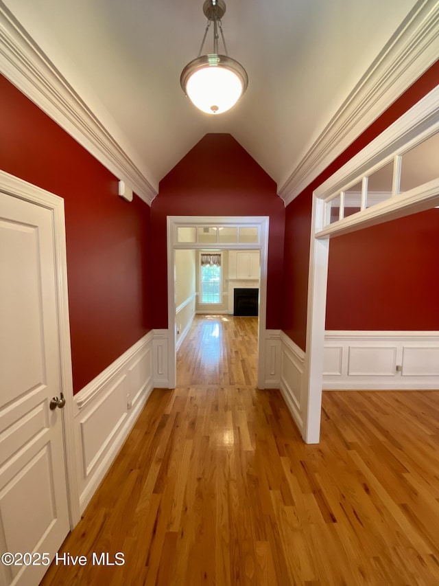 corridor featuring vaulted ceiling, wainscoting, a decorative wall, light wood finished floors, and crown molding