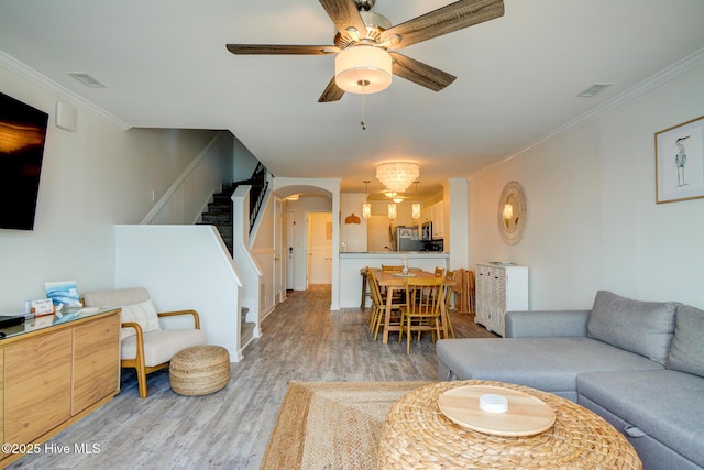 living room featuring stairway, light wood-style flooring, crown molding, arched walkways, and ceiling fan