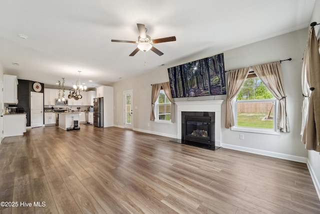 unfurnished living room featuring ceiling fan, a chandelier, a glass covered fireplace, dark wood finished floors, and baseboards